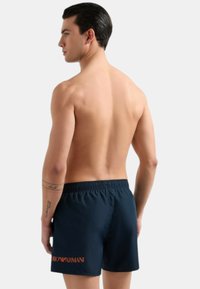 Pantaloni da bagno blu navy con cintura elastica, dotati del logo arancione "Armani" sul retro. Tessuto liscio e leggero.