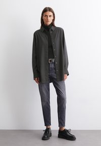 Ylipitkä tummanvihreä nappipaita mustan turtleneckin päällä, yhdistettynä harmaisiin slim-fit-farkkuihin ja mustiin nauhakengiin.
