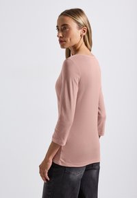 Femme aux cheveux blonds portant un haut à manches longues rose pâle et un jean noir délavé, debout légèrement de profil sur un fond blanc.