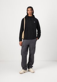 Felpa nera in pile con tasca kangaroo, pantaloni cargo grigi con tasche laterali e sneakers beige. Design semplice, vestibilità rilassata, nessun motivo visibile.