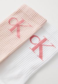 Kokvilnas ribotas zeķes rozā un baltā krāsā, katra ar lielu izšūtu "CK" logotipu kontrastējošā rozā.