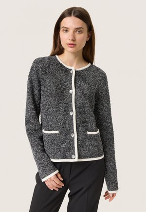 Black and white textured cardigan with round neckline, front pockets, and shiny buttons. Trimmed in white, featuring a fitted silhouette.