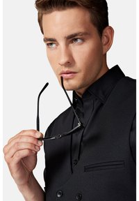 Czarna, teksturowana kamizelka z zapięciem na guziki, noszona na ciemnej koszuli. Ręka trzyma czarne okulary w pobliżu brody, podkreślając kieszeń kamizelki.