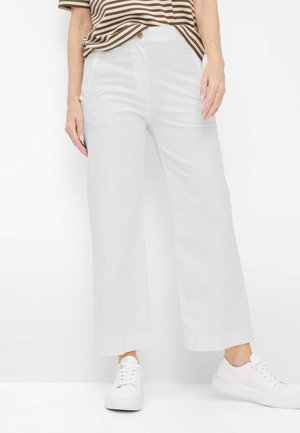 Kvinde iført hvide bukser med høj talje og vide ben, brunstribet skjorte og hvide sneakers, stående mod en hvid baggrund.