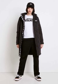 Veste longue noire matelassée avec capuche, t-shirt graphique blanc, pantalon noir et baskets à carreaux noir et blanc, casquette noire assortie.