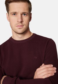 Bordowy, teksturowany sweter z okrągłym dekoltem i subtelnym logo na lewej piersi, noszony przez osobę z założonymi rękami.