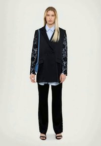 Black blazer with lace sleeves, layered over a light blue shirt. Paired with black trousers and sandals. Blue shoulder bag. Neutral background.