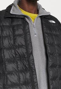 Svart vadderad puffer-väst över en grå fleece-pullover med gul kringhals, med dragkedja fram och kontrasterande sömmar.