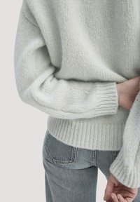 Hellgrauer Strickpullover mit weicher Textur und geripptem Saum, kombiniert mit mittelgrauen Jeans. Der Pullover hat eine lockere Passform mit weiten Ärmeln.