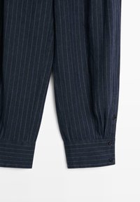 Marineblauwe broek met fijne strepen, omgeslagen zoom en knoopdetails op het onderste deel van de pijpen, afgebeeld tegen een witte achtergrond.