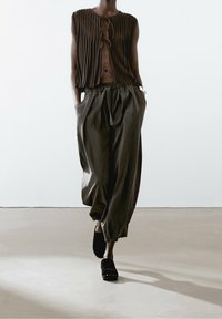 Brown pleated sleeveless top with tie detail, paired with loose gray trousers. Black clogs with metallic accents complete the outfit.