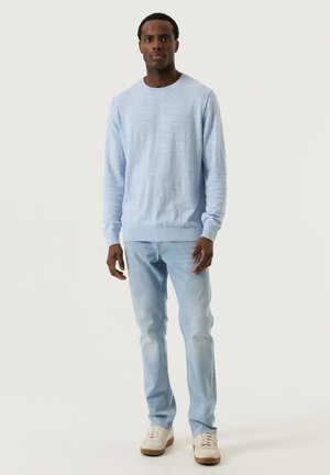 Mann steht in einem hellblauen Pullover, hellblauen Jeans und beigen Turnschuhen vor einem schlichten weißen Hintergrund.