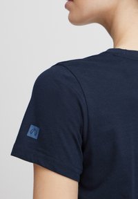 Camiseta de algodón azul marino, de cuello redondo, con mangas cortas y un pequeño parche con logotipo azul en el hombro izquierdo. Textura suave.