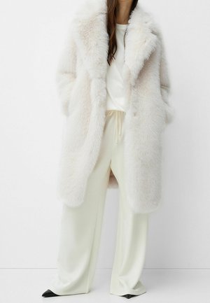 Femme portant un long manteau en fourrure blanche duveteuse par-dessus un haut blanc et un pantalon large crème avec des chaussures noires pointues, debout contre un mur blanc.