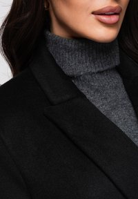 Svart yllekappa med en strukturerad krage över en texturerad mörkgrå polotröja. Närbild som framhäver tyg- och designdetaljer.