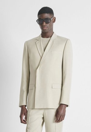 Man draagt een oversized beige pak en trui met grote zwarte zonnebril, staand tegen een effen witte achtergrond.