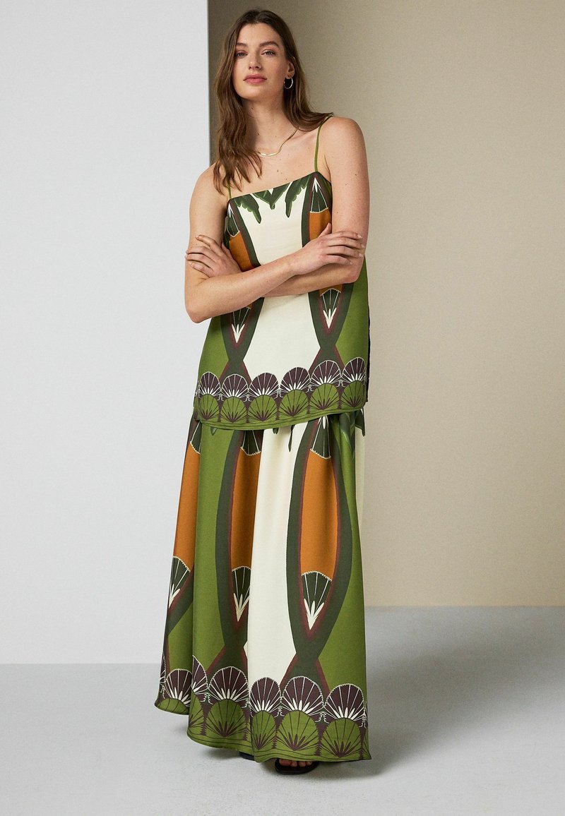 Femme debout, bras croisés, portant un top sans manches à motifs verts, orange et crème et un pantalon large assorti.