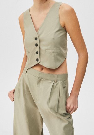 Frau trägt eine ärmellose beige Weste mit Knöpfen und hochtaillierte beige plissierte Hosen mit Händen in den Taschen.