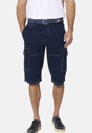 Mann trägt dunkelblaue Cargo-Shorts mit großen Seitentaschen, geflochtenen Gürtel, weißes T-Shirt und schwarze Sneakers auf schlichtem Hintergrund.