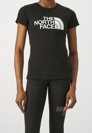 Persoon draagt een nauwsluitend zwart T-shirt met wit "The North Face"-logo en zwarte legging met gedeeltelijke logotekst op de dij.