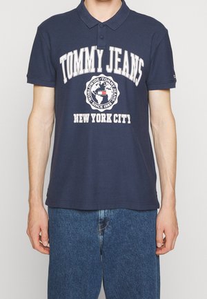 Mężczyzna w granatowej koszulce polo Tommy Jeans z białym napisem „Tommy Jeans New York City” i logo globu, w zestawieniu z niebieskimi dżinsami denim.