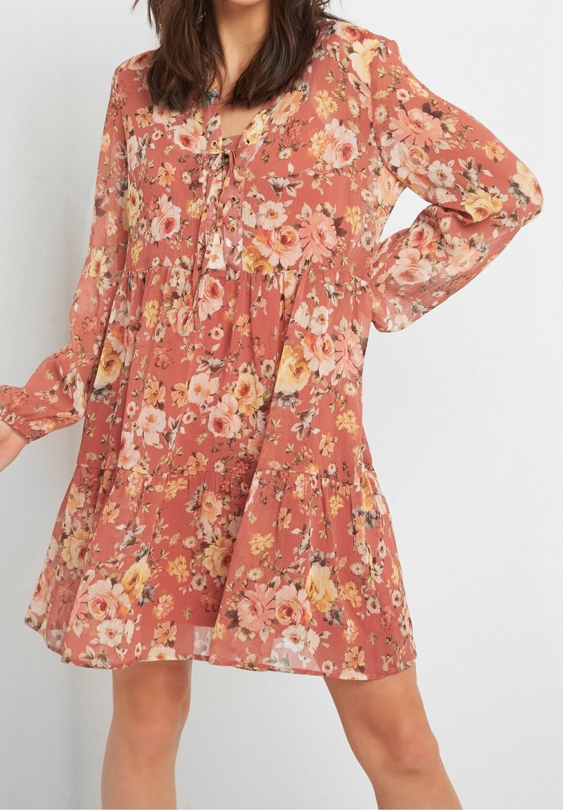 Femme portant une robe corail fluide à manches longues avec un imprimé floral et un col à lacets, se tenant devant un fond clair uni.