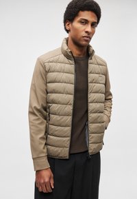 Veste matelassée beige avec un tissu lisse sur la partie supérieure et une section inférieure matelassée, dotée d'un col montant, d'une fermeture éclair avant et d'un patch logo. Pull marron texturé en dessous.