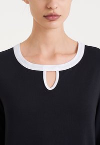 Top blu navy con colletto rotondo bianco e dettaglio a oblò sul davanti. Tessuto morbido, design semplice, forma aderente, contrasto di colori classico.