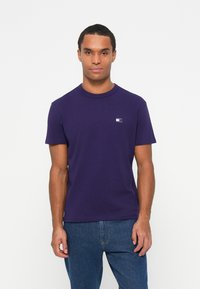 T-shirt di cotone viola con collo rotondo, maniche corte e un piccolo logo sul lato sinistro del petto, abbinato a jeans denim blu.