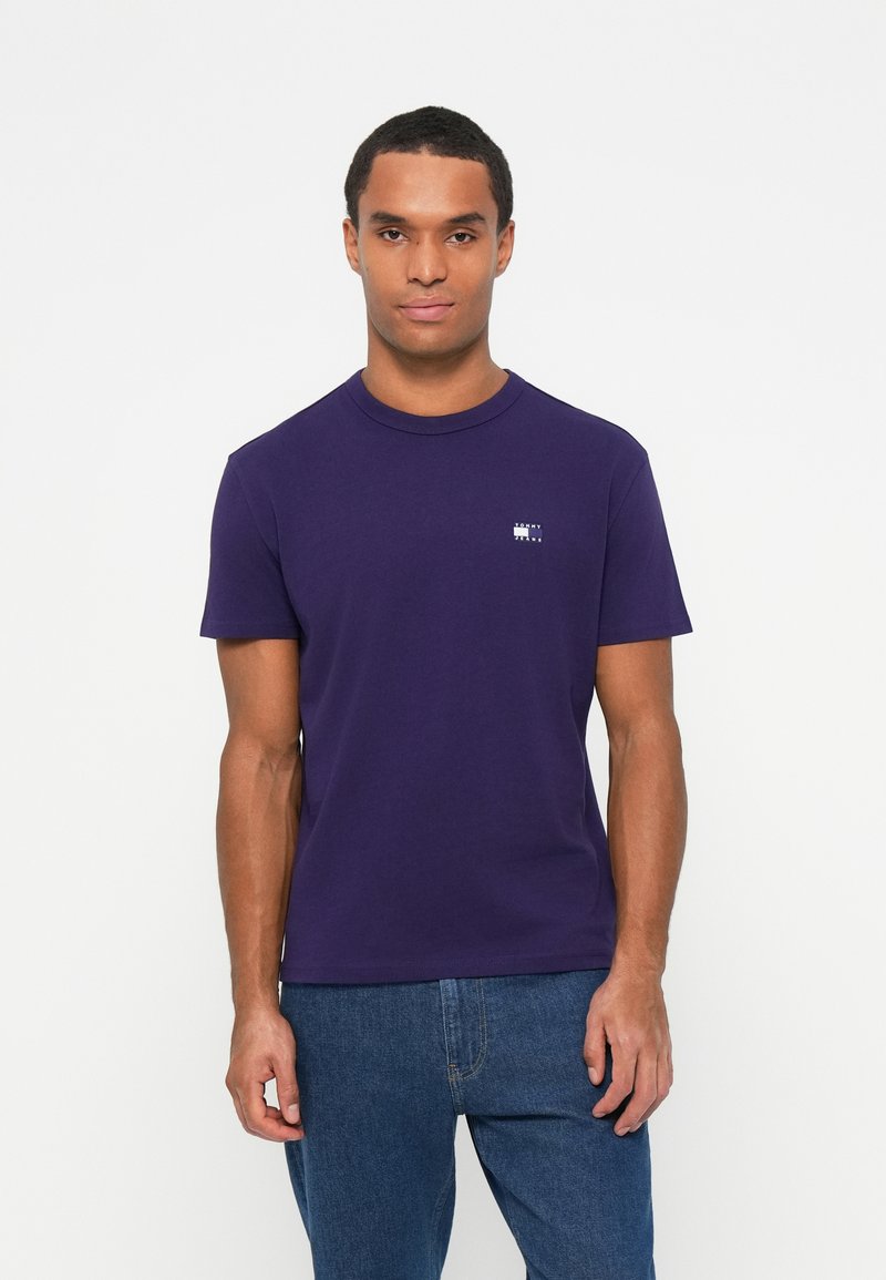 T-shirt di cotone viola con collo rotondo, maniche corte e un piccolo logo sul lato sinistro del petto, abbinato a jeans denim blu.