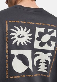 Γκρι βαμβακερό t-shirt με τέσσερα μεγάλα λευκά γραφικά σχέδια: ήλιο, μάτι, ανθό και παλάμη, με πορτοκαλί κείμενο που περιβάλλει το σχέδιο.