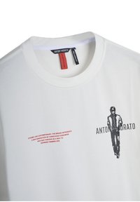 Camiseta blanca de algodón de manga corta con un diseño gráfico negro de una figura y texto rojo en el lado izquierdo. Etiquetas de marca visibles en el cuello.