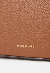 Cerca de cuero marrón con textura de piedras, presentando el logotipo grabado "MICHAEL KORS" en dorado. Superficie texturizada con un acabado ligeramente brillante.