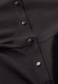 Czarna koszula z gładkiej tkaniny, z pięcioma czarnymi guzikami ułożonymi wzdłuż środka oraz przeszyciami w okolicach ramion.