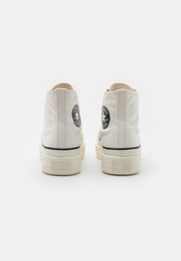 White canvas high-top sneakers with black rubber accents and circular logo patch on the heel. Textured upper and thick sole.