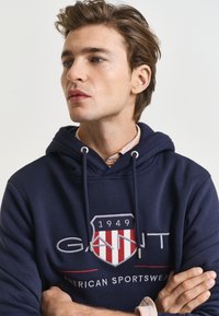 Felpa in cotone blu navy con cappuccio regolabile, presenta un logo ricamato e la data 1949 in rosso e bianco sul davanti. Texture liscia.