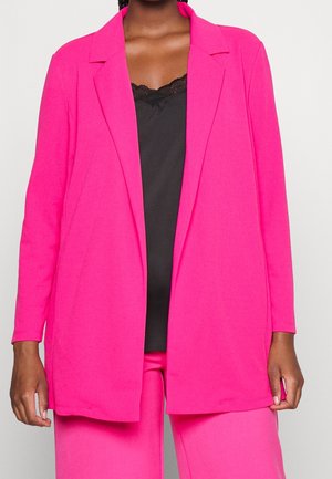 Mujer con blazer y pantalones color rosa brillante y una blusa negra con encaje, mostrada desde los hombros hasta la mitad del muslo sobre un fondo liso.