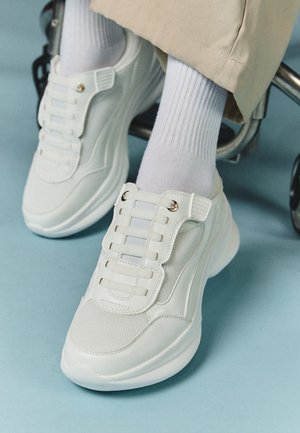 Witte sneakers met een mesh materiaal, rubberen zool, vetersloze uitvoering met elastische banden en gouden accenten.