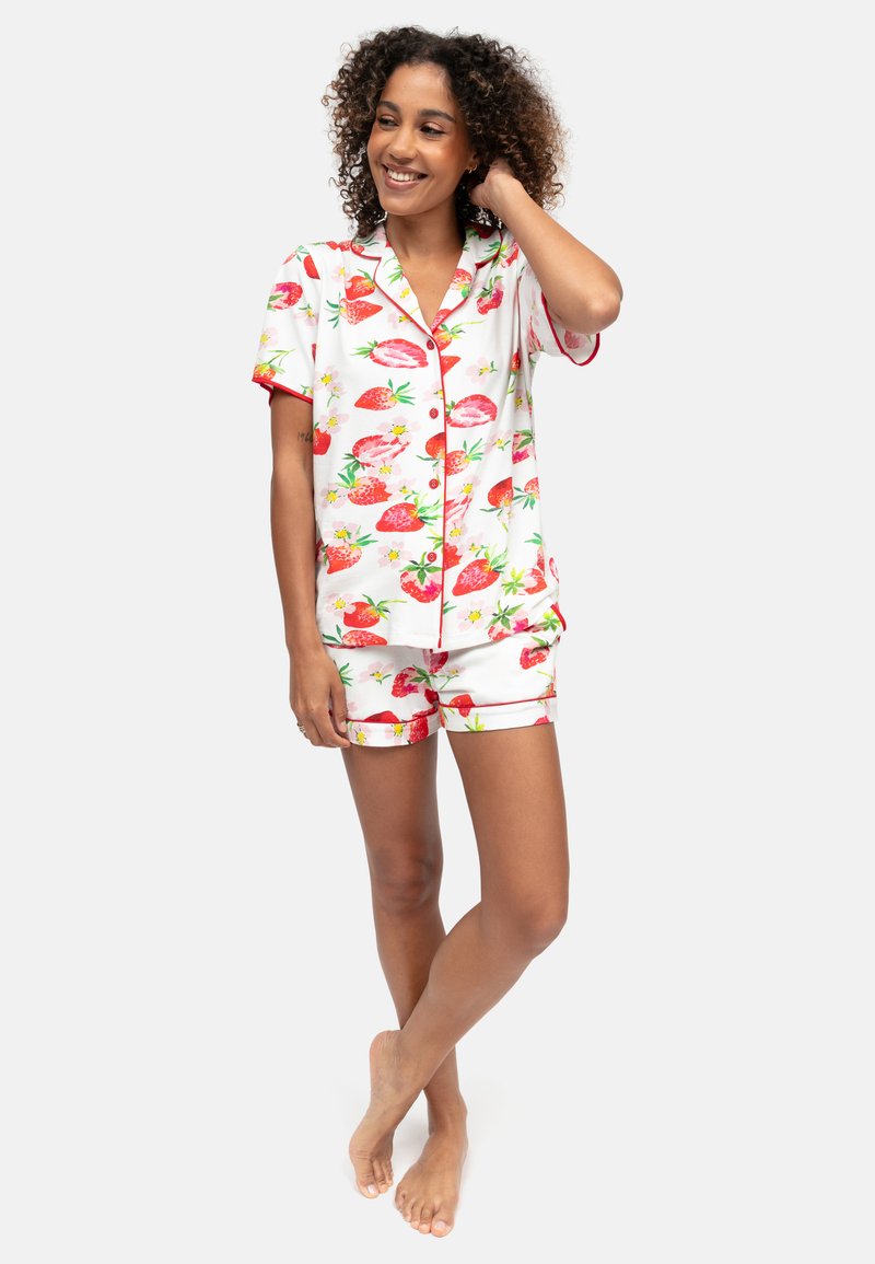 Uśmiechnięta kobieta o kręconych włosach, ubrana w biały zestaw piżamowy z krótkim rękawem na guziki i szorty z czerwonym nadrukiem truskawek, stojąca boso.