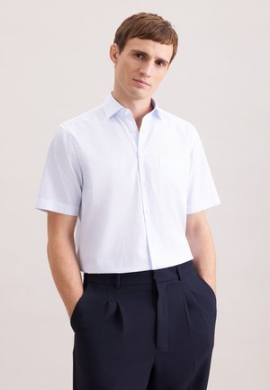 Mand iført en kortærmet hvid skjorte med knapper og mørke plisserede bukser, stående med hænderne i lommerne mod en ensfarvet baggrund.