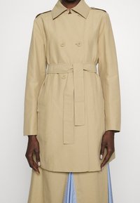 Trench-coat beige avec une coupe croisée à double boutonnage, une ceinture à la taille, des pattes d'épaule et des poches avant. Tissu lisse avec une coupe ajustée.