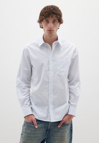 Camicia bianca con strisce blu sottili, dotata di taschino sul petto e maniche lunghe, indossata con jeans in denim blu.