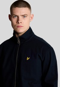 Giacca zip-up blu navy con colletto alto, realizzata in un materiale liscio con un piccolo logo giallo in evidenza sul petto.