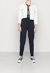Giacca di jeans bianca accorciata su una camicia a stampa floreale, abbinata a pantaloni tapered blu scuro e sneakers bianco e nere. Texture lisce.