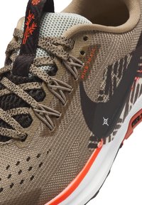 Zapatilla de trail running con una parte superior de malla marrón texturizada, detalles en negro y naranja, y una suela robusta. Presenta un logo destacado de Nike.