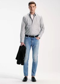 Chemise grise à boutons, t-shirt blanc, jeans bleu clair, ceinture noire, veste noire à la main et chaussures noires. Le modèle se tient devant un fond uni.