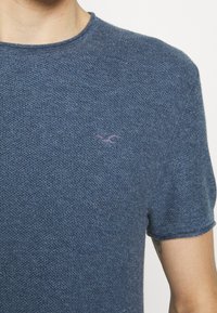 Modré pletené tričko s krátkým rukávem, s texturovaným vzorem a malým vyšitým logem na hrudi. Kulatý výstřih s vroubkovaným lemem.