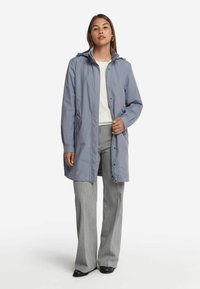 Ljusgrå, vattentät långkappa med en åtsittande huva, framzipper och sidofickor. Bärs över en vit topp och grå byxor.