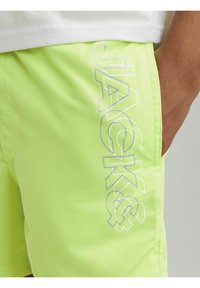 Bañador verde neón con una textura suave, que presenta un diseño gráfico blanco "JACKS&" en el lateral y un bolsillo lateral.