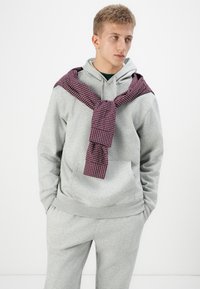 Haut à capuche gris avec poche kangourou, associé à un pantalon de survêtement assorti. Une chemise à carreaux bordeaux est nouée autour du cou. Design simple et décontracté.